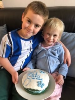 ed girl with her brother sat on the sofa looking at the camera holding their blue and white ED cake they baked for fundraising 