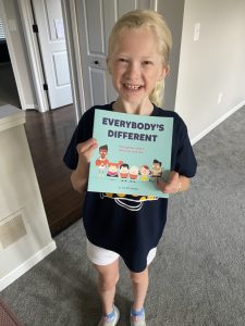 a blonde little girl with EEC standing in frot of the camera holding a copy of the ED Society book everybody's different