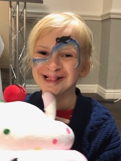 Elsie is a 6 year old girl with blonde hair tied back, smiling into the camera cuddling a pink pig teddy. She has her face painted with a wave and a penguin swimming